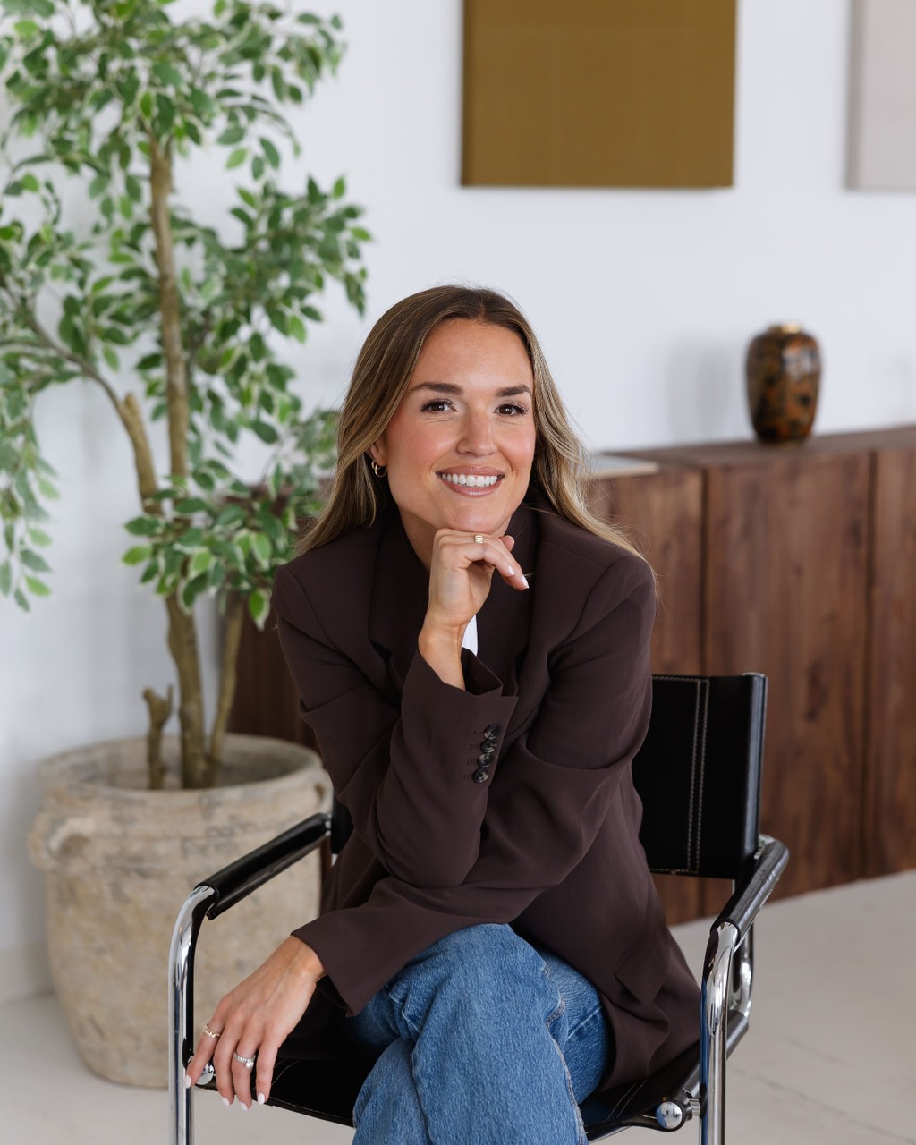 Olivia, founder of The List, smiling in a chocolate blazer and jeans seated on a black chair