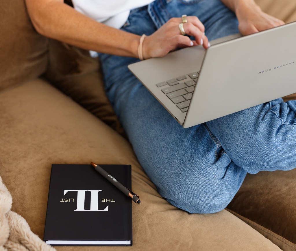 A woman on a brown velvet sofa with a laptop and a branded The List notebook